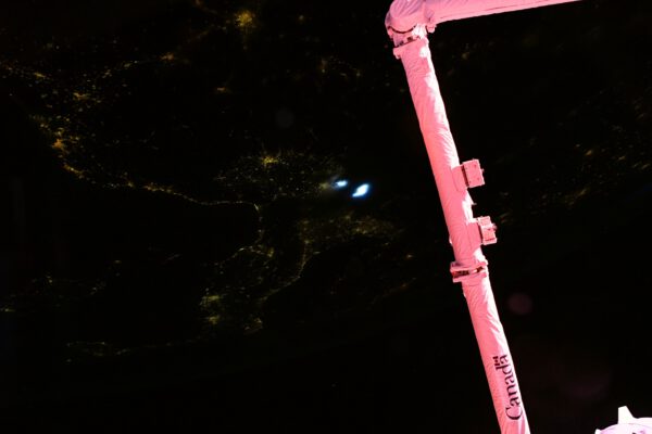 Zde už je v pozadí vidět světla měst v noci, přesto na Canadarm-2 ještě dopadají poslední paprsky Slunce. Obarvily ji krásně do červena až růžova. Nechybí ani dva světlé zdroje světla od blesků v bouřkovém oblaku. Zdroj: Flickr.com