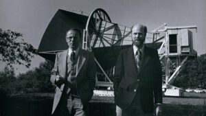 Arno Penzias (vlevo) a Robert Woodrow Wilson (vpravo). Za nimi je anténa s jejíž pomocí objevili reliktní záření.