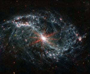 NGC 7496, jak ji viděl přístroj MIRI. Kromě samotného jádra galaxie, které je dokonce tak jasné, že u něj vidíme i difrakční hroty, si povšimněte také tmavšího okolí snímku. Zde totiž najdeme i několik objektů hlubšího vesmíru zachycených je tak mimochodem.