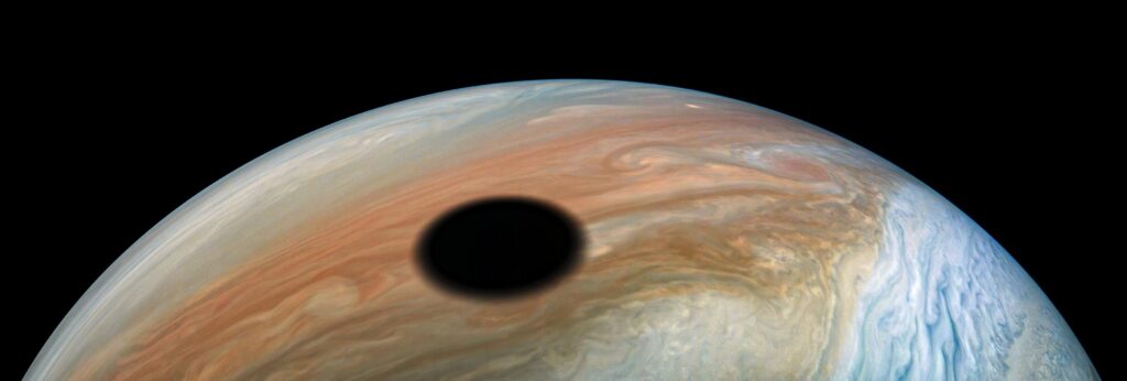 Na snímku z 11. září 2019 pořízeném americkou sondou Juno je stín měsíce Io, který putuje po svrchní vrstvě oblačnosti Jupitera. Náš neexistující pozorovatel nacházející se v tomto temném kruhu o průměru přes 3500 km by zažíval úplné zatmění Slunce.